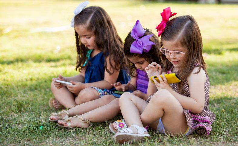 Cute Little Girls Playing With Their Smartphones Ignoring One Another In A Park - Social Media And Technology Effect